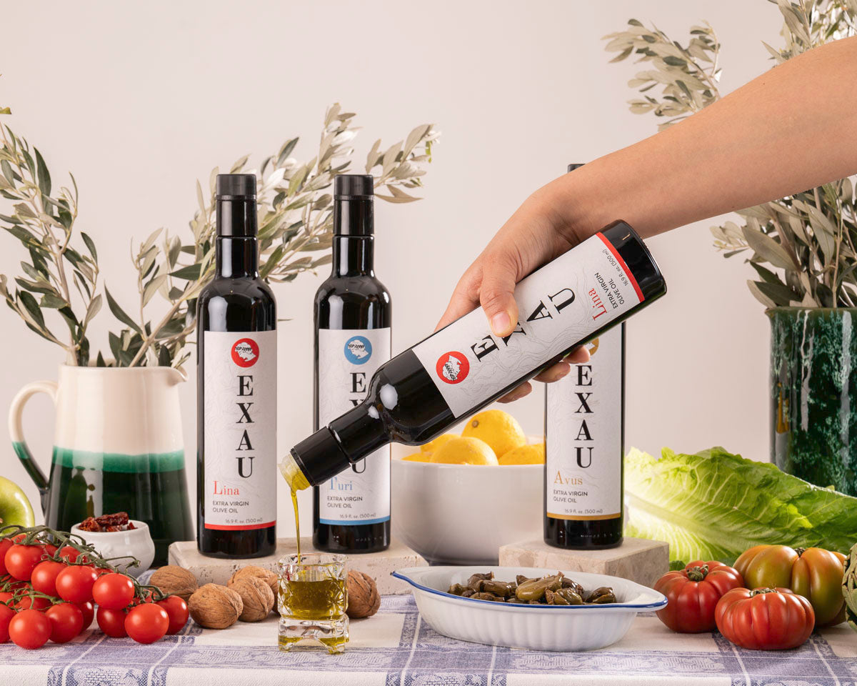 Four bottles of EXAU olive oil displayed with a backdrop of olive branches and a variety of fruits and vegetables on a table with a person pouring oil into a small container